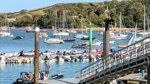 A marina with various boats docked, a pedestrian bridge, and a hilly landscape in the background. - Firm Anchor (Salcombe)