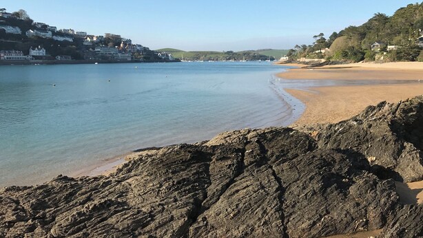 A coastal beach with rocky foreground, calm waters, and buildings in the background. - Firm Anchor (Salcombe)