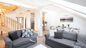 A spacious living area with two grey sofas, a wooden staircase, a kitchen in the background, and a dining area with a table and chairs. - 150m² 4BR Duplex Near Metro & Prague Center (Hlavní mesto Praha)