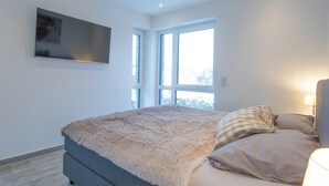 Une chambre à coucher avec un lit, un téléviseur fixé au mur et de grandes fenêtres.