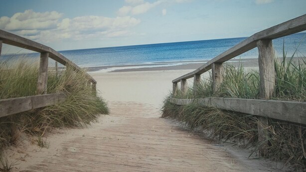 Une promenade en bois mène à une plage de sable, avec l’océan en toile de fond.