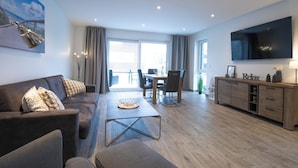 Un salon moderne avec un canapé, une table basse et un téléviseur. Il y a également un coin repas avec des chaises et une table. La pièce est dotée d’un parquet et de grandes fenêtres.