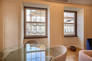 A room with a glass table, two chairs, a small rug, and two windows with a view of buildings.