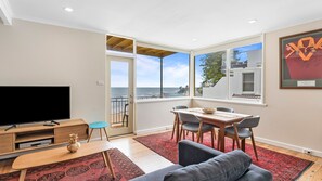 A modern living room with a large TV, a dining area with chairs and a table, and a balcony with a view of the sea.