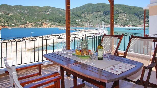 A wooden table with a glass of wine and a plate of fruit, overlooking a coastal area with boats and hills.