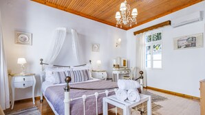 A bedroom with a four-poster bed, a bedside table with a lamp, a small table with towels, a wooden ceiling, and a window with curtains.