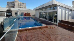 Una piscina en la azotea con barandillas de vidrio, una terraza de madera y un edificio moderno con grandes ventanales y fachada blanca.