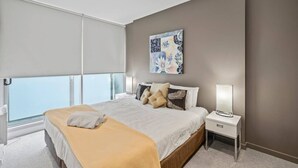 Una habitación de hotel moderna con una cama grande, una mesita de noche con lámpara y vistas a la ciudad a través de ventanales de gran altura.