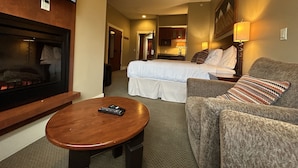 A hotel room with a TV mounted on the wall, a bed with white bedding, a gray sofa with a patterned throw pillow, and a wooden coffee table.