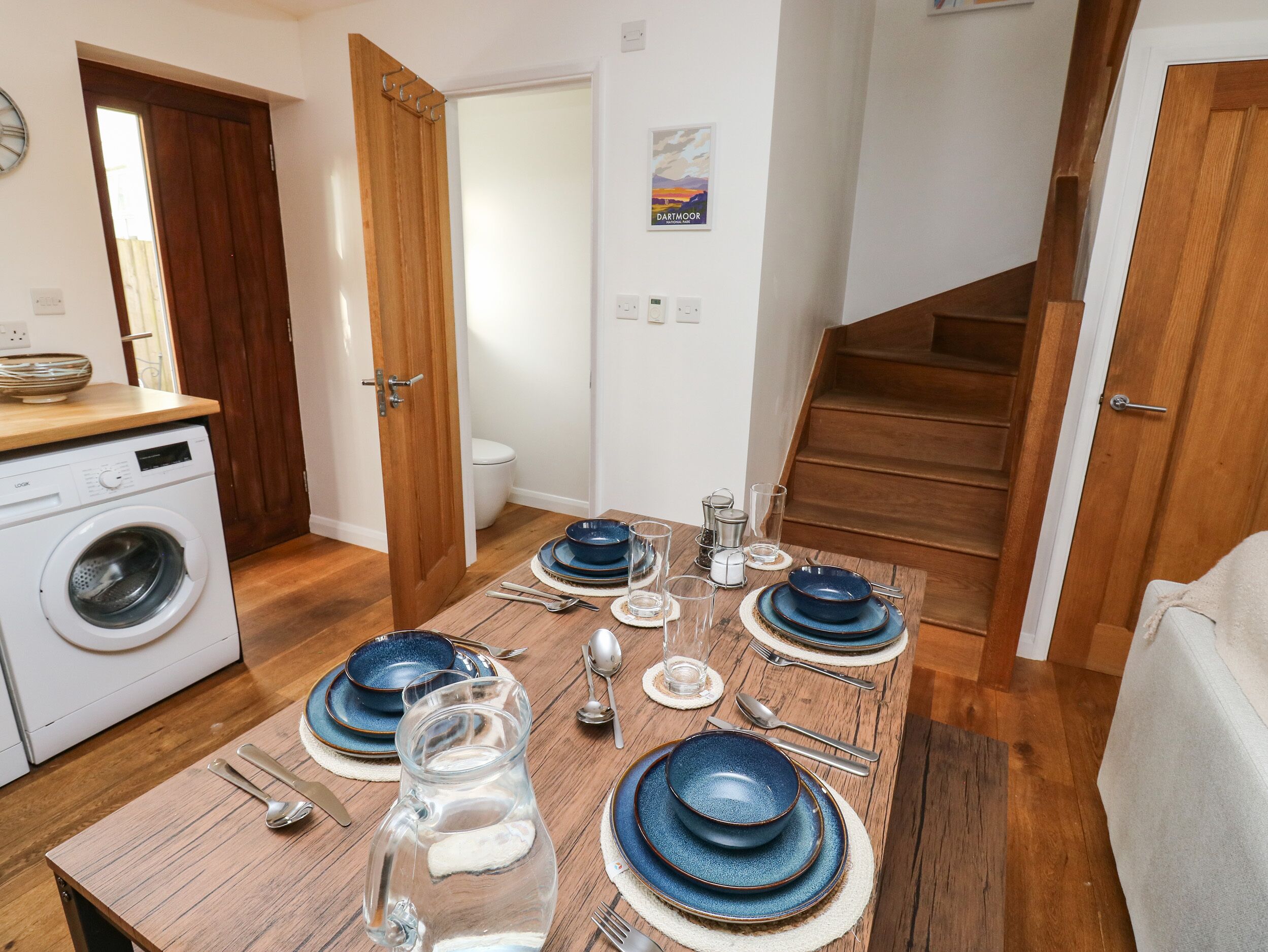Comedor con mesa de madera dispuesta para cuatro personas, lavadora y escalera que sube a otro nivel.