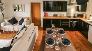 A modern kitchen with black cabinets, a wooden dining table set for four, and a cozy living area with a sofa and a framed picture on the wall. - Little Rondeval (Totnes)