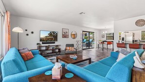 Un salon spacieux avec un grand canapé bleu, une table basse en bois et un téléviseur. On y trouve également un coin repas avec des chaises rouges et une cuisine visible au fond.