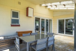 Eine überdachte Terrasse mit einem Glastisch und Stühlen, einer Holzbank und bodentiefen Glasschiebetüren zu einem Außenbereich.