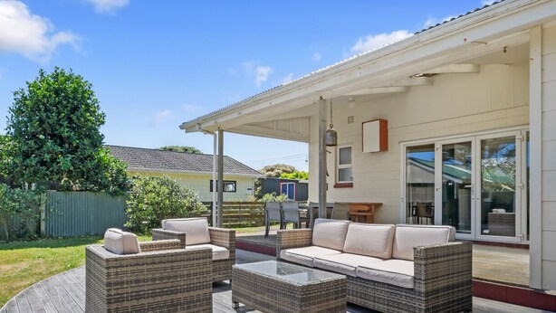 Um pátio externo com móveis de vime e mesa de vidro, área coberta com porta de correr e vista para uma área residencial.