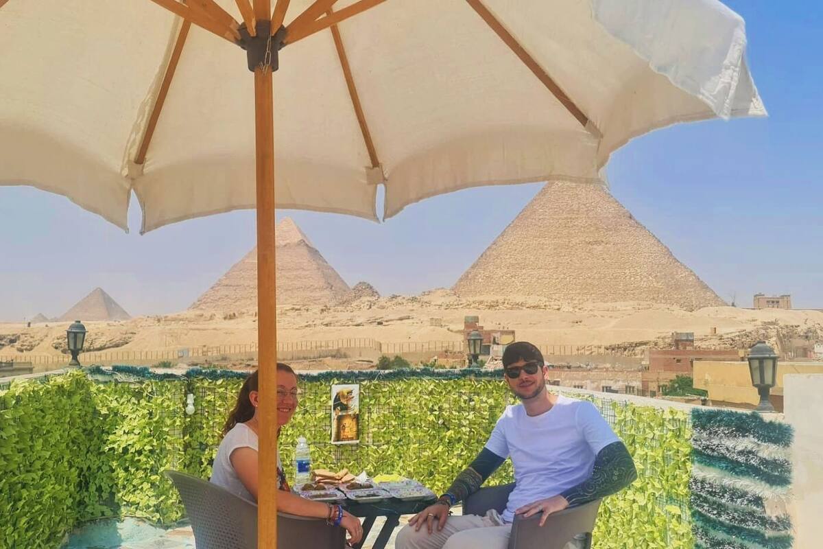 A dining area with a table and chairs under a large umbrella, with a view of pyramids in the background.