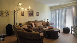 Una sala de estar con un sofá modular, una mesa de centro y una lámpara de pie. Se aprecia un ventanal con persianas, un televisor y una chimenea.