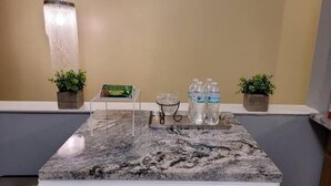 Una encimera de cocina con cubierta de mármol, dos plantas en macetas, una bandeja con botellas de agua y una lámpara colgante.