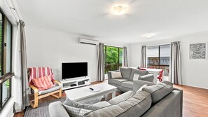 A modern living room with a flat-screen TV, grey sofa, and a dining area with red chairs.
