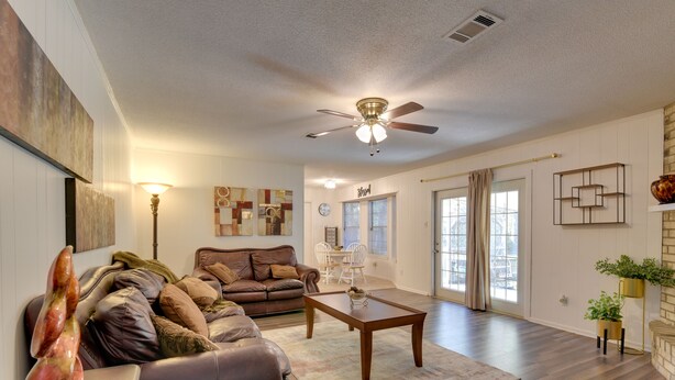 A spacious living room with a brown leather sofa, a wooden coffee table, and a ceiling fan. There is a dining area with a white table and chairs, and a large window with curtains. - Beaumont Rental Home ~ 2 Mi to Gulf Terrace Park! (Beaumont)