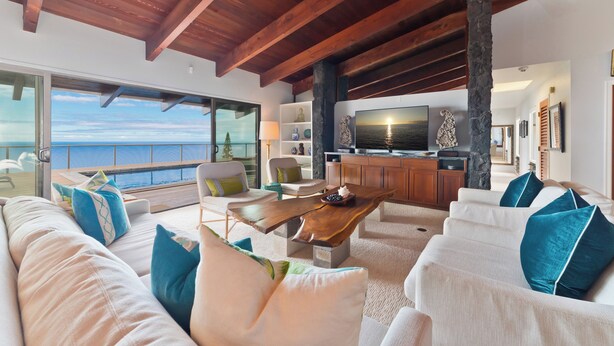 Een ruime woonkamer met een houten plafond, een grote houten salontafel, witte banken met blauwe kussens en uitzicht op de oceaan door grote ramen.