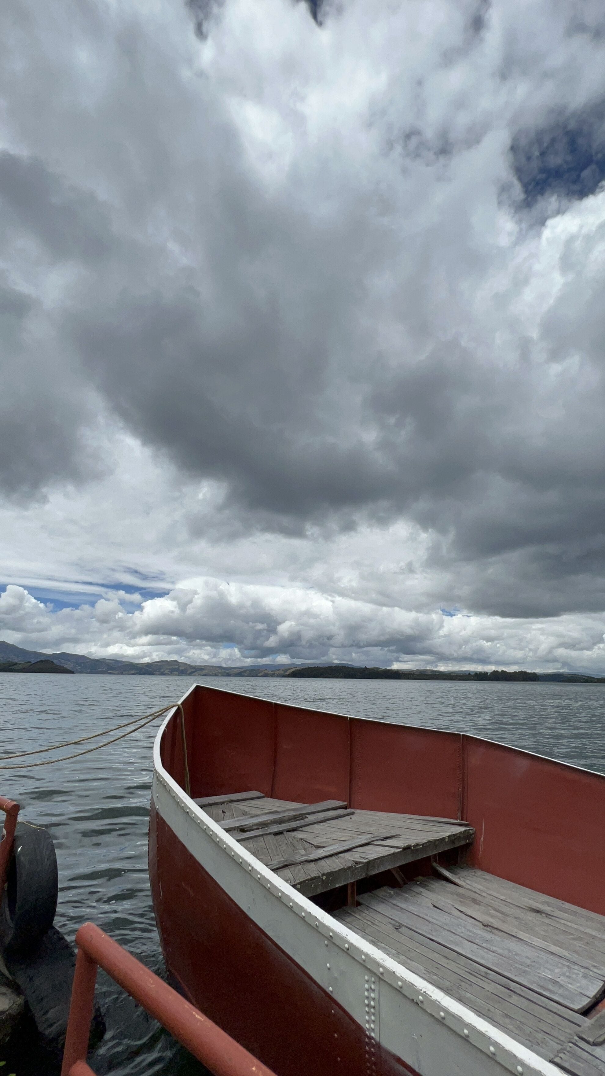 Um barco com interior vermelho e deck de madeira, atracado em águas calmas sob um céu nublado.