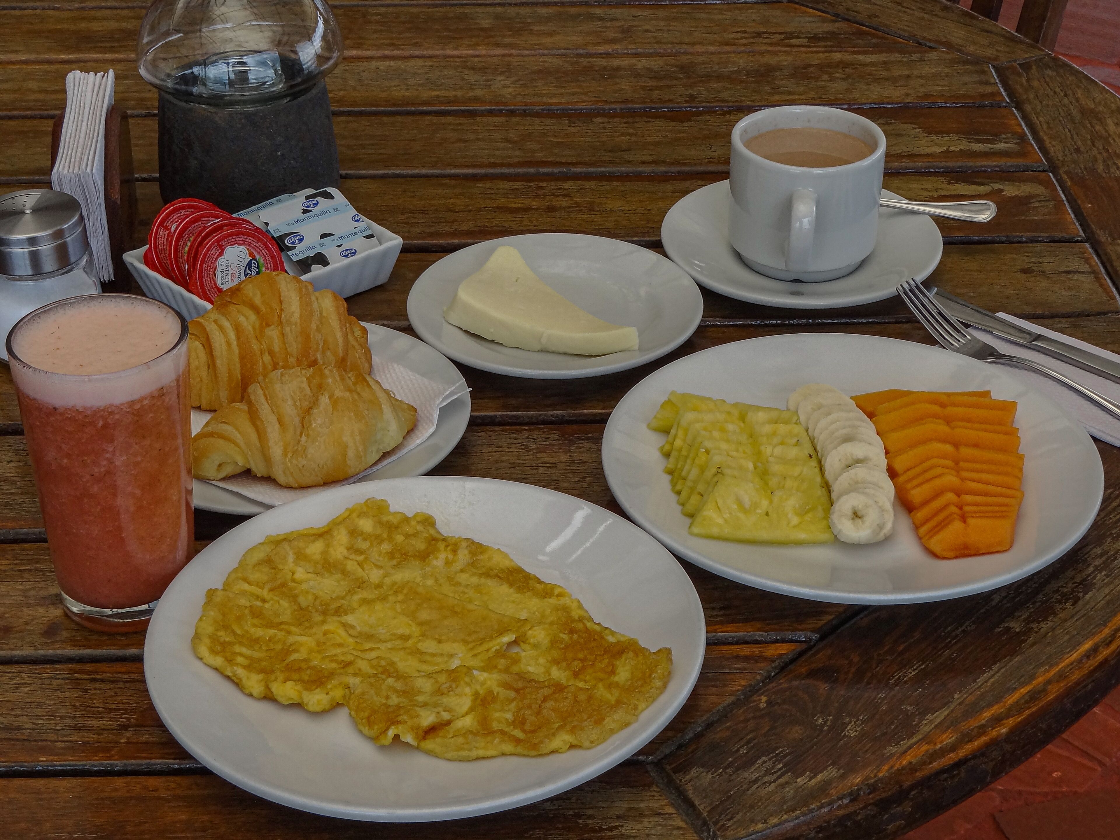 A breakfast table with croissants, a smoothie, a plate of fruit, a plate of eggs, a cup of coffee, and a bottle of milk.