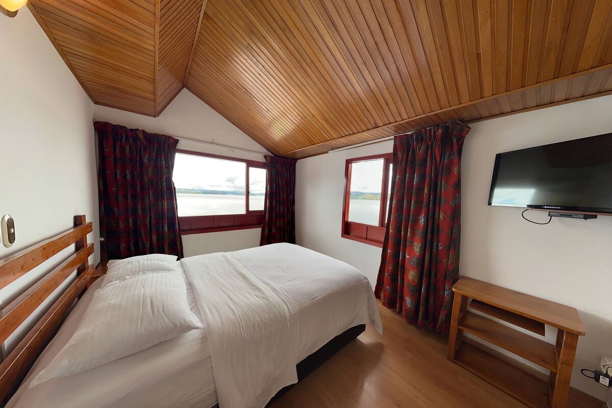 Un dormitorio con techo de madera, una cama, un escritorio, un televisor y ventanas con cortinas.