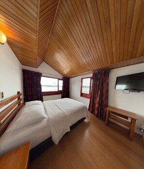 A bedroom with a wooden ceiling, a bed, a desk, a TV, and windows with curtains.