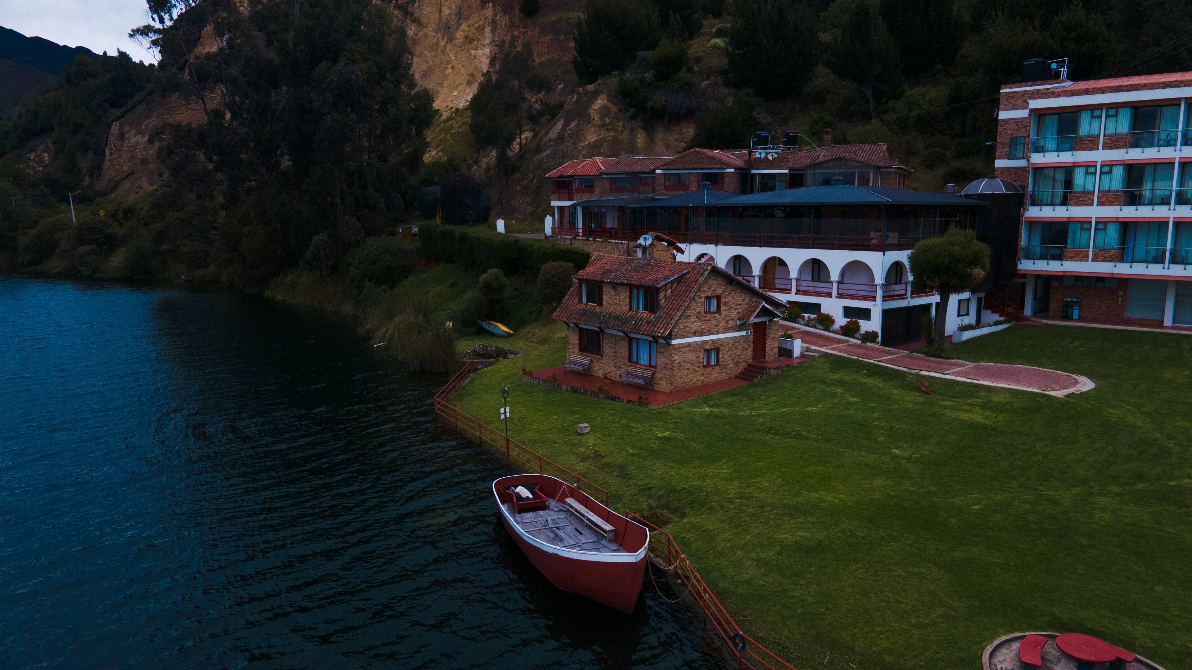 Vista aérea de una propiedad junto al lago con un bote amarrado cerca del agua, un edificio grande con varios balcones y una casa más pequeña con techo rojo.