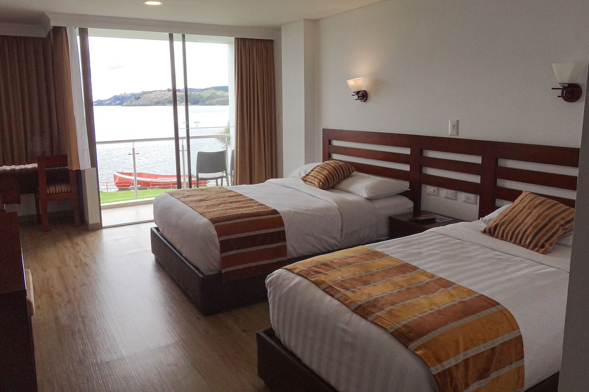 Habitación de hotel con dos camas, un escritorio y vistas a una masa de agua a través de amplios ventanales.
