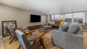 A living room with a flat-screen TV, grey sofas, wooden chairs, a coffee table, and a large window with a city view.