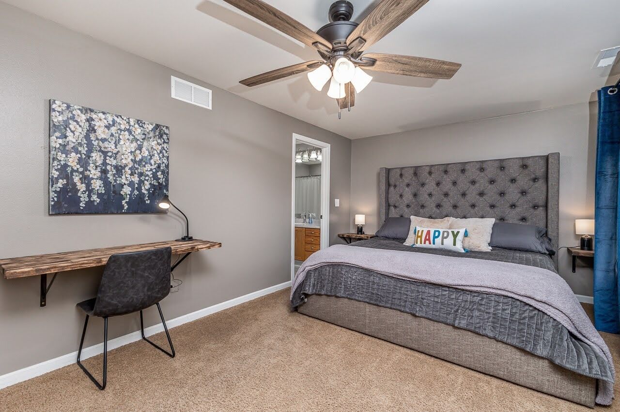 A bedroom with a bed, a desk, a chair, a ceiling fan, and a wall-mounted artwork.