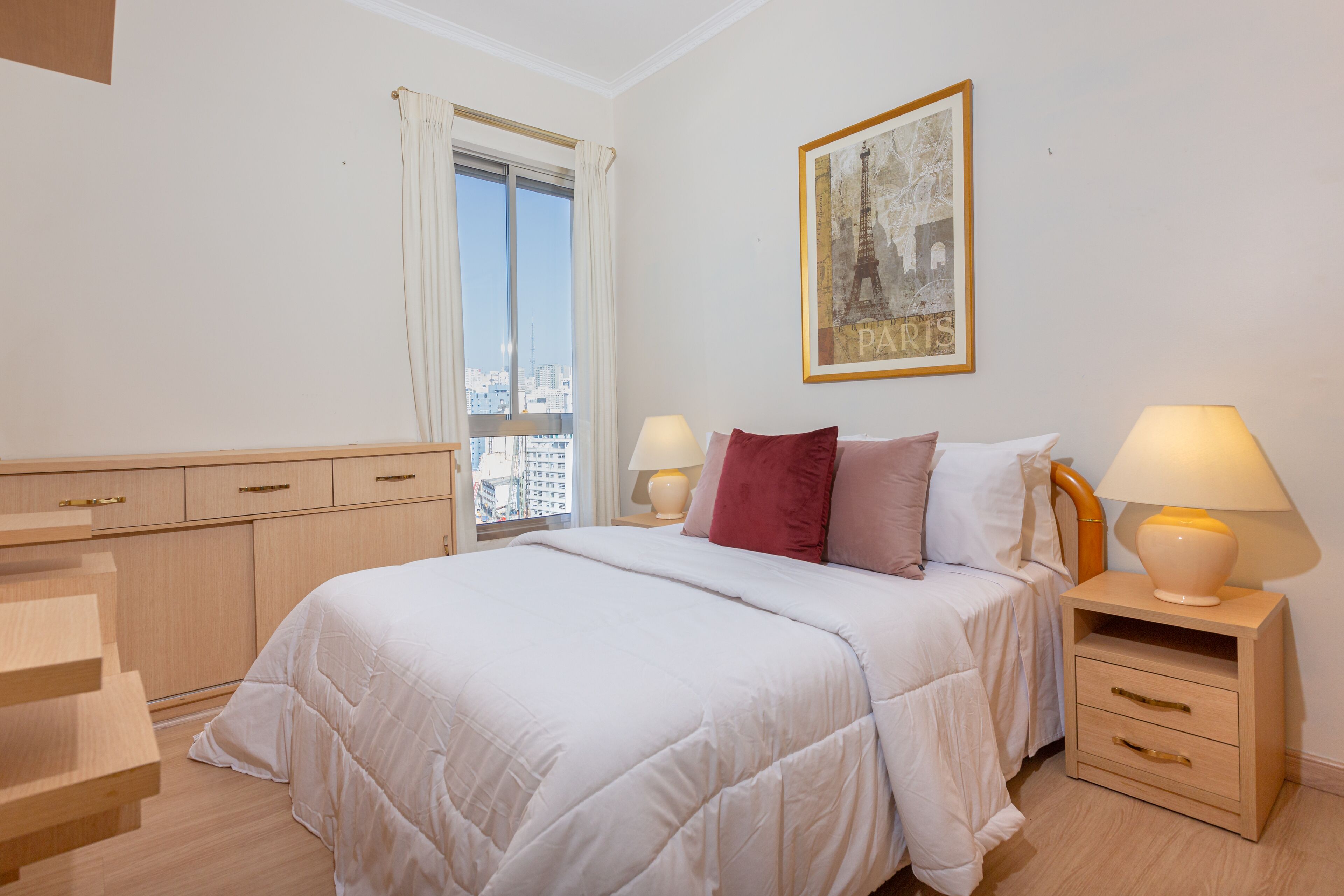 A hotel room with a bed, bedside tables with lamps, a window with curtains, a framed picture of the Eiffel Tower, and a view of the cityscape.