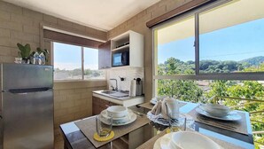 Cocina moderna con refrigerador, microondas y pileta. Hay una mesa de comedor dispuesta con platos y vasos. La habitación tiene ventanas amplias con vistas a zonas verdes y edificios.