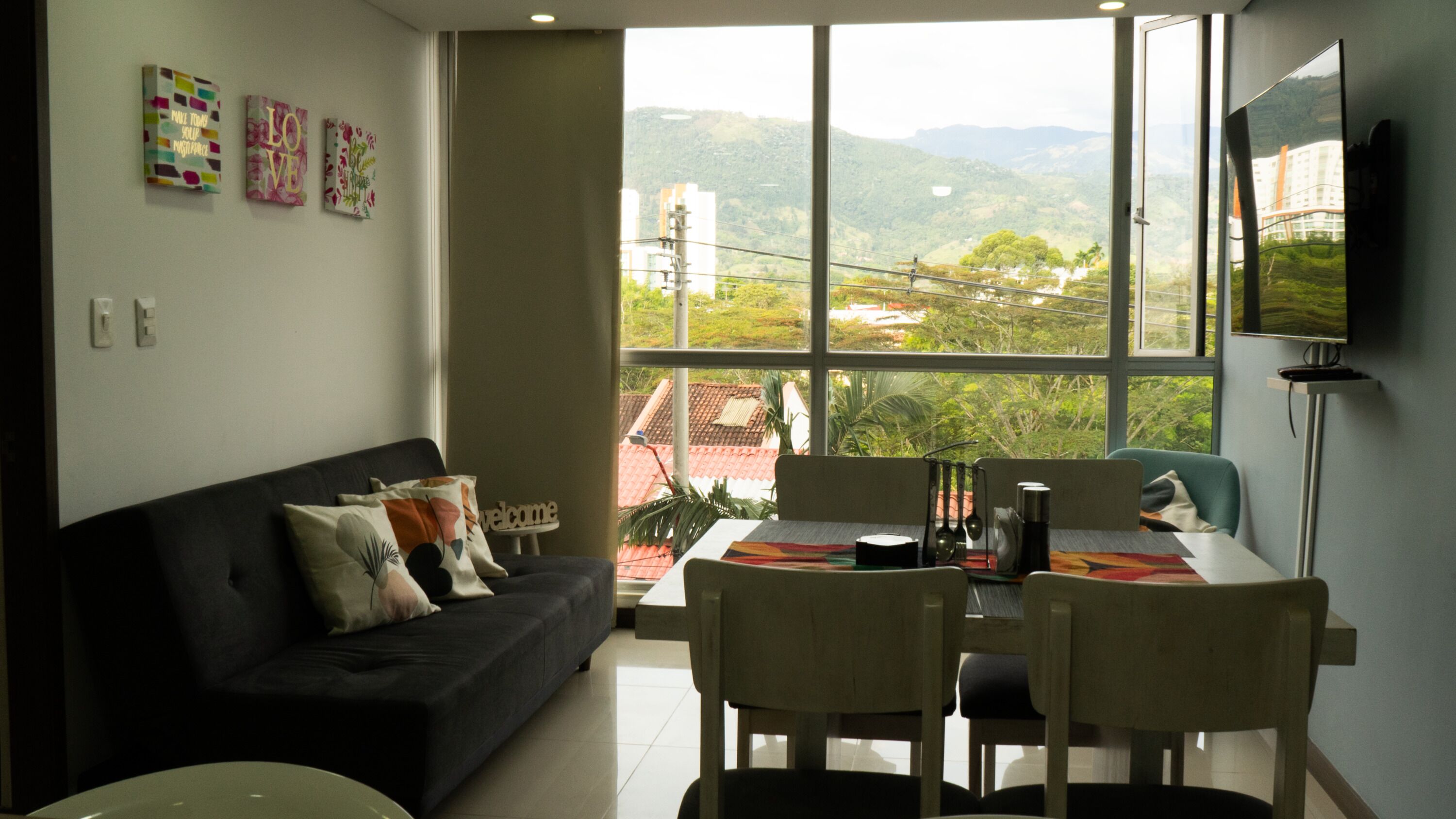 A modern living room with a black sofa, a dining table with chairs, and a large window offering a view of a hilly landscape.