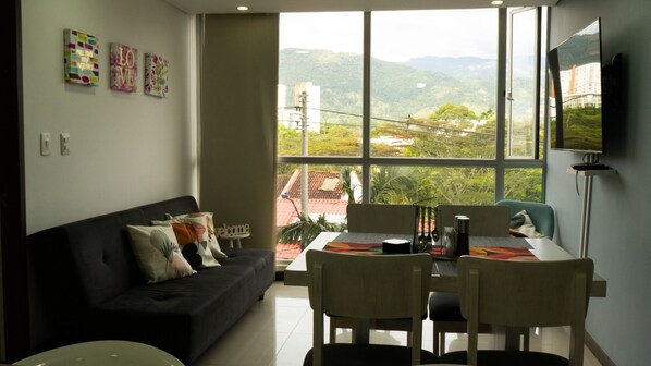 A modern living room with a black sofa, a dining table with chairs, and a large window offering a view of a hilly landscape.