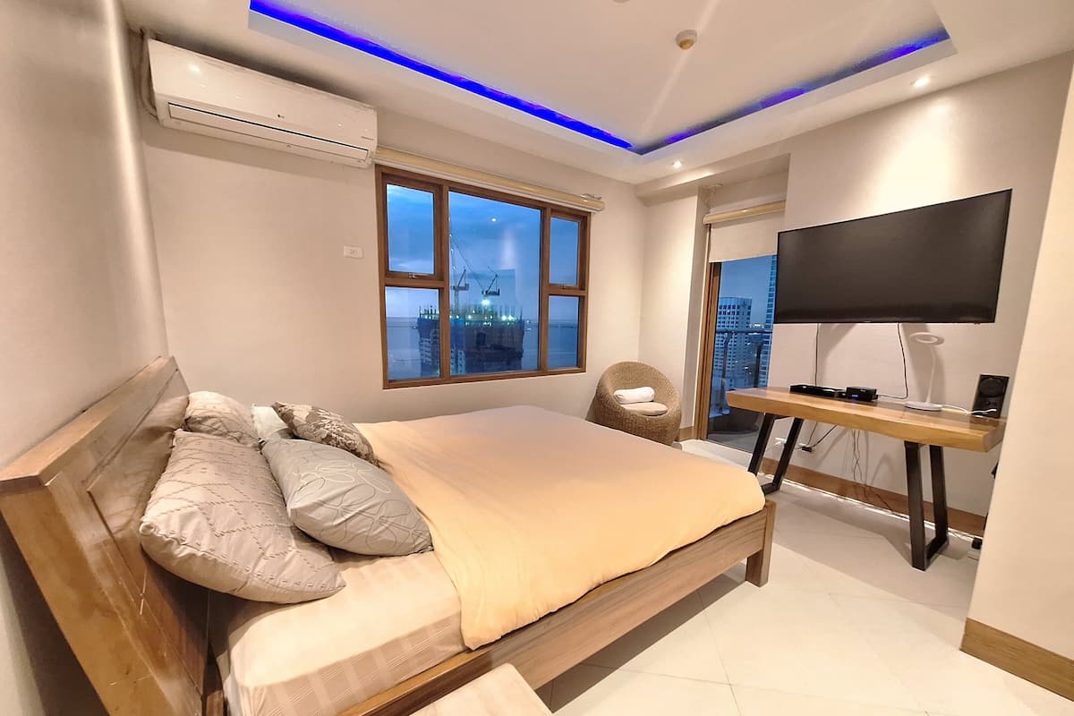 A modern bedroom with a large bed, a TV, and a city view.