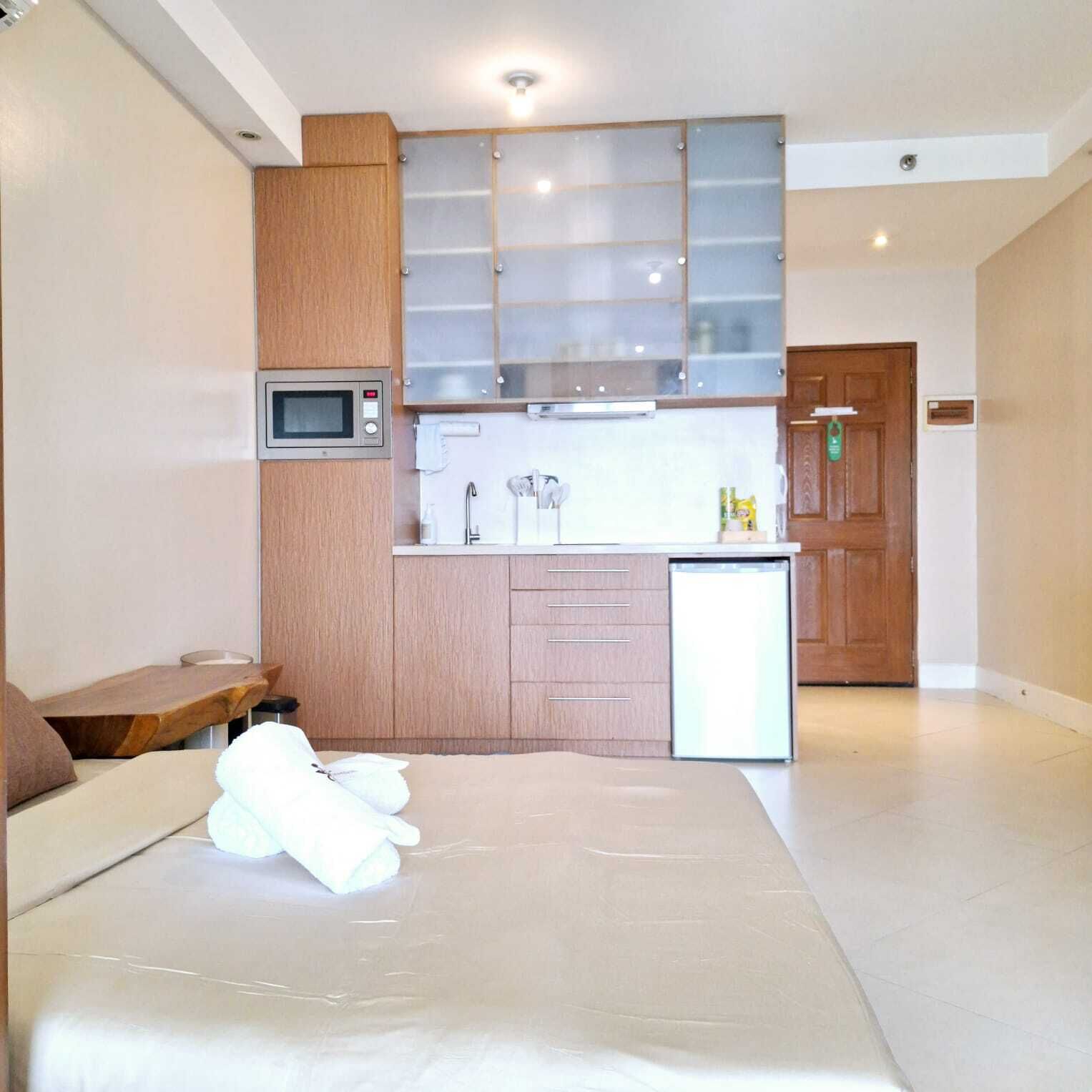 A compact bedroom with a bed, a small bedside table, and a built-in kitchenette with a microwave, sink, and storage cabinets.
