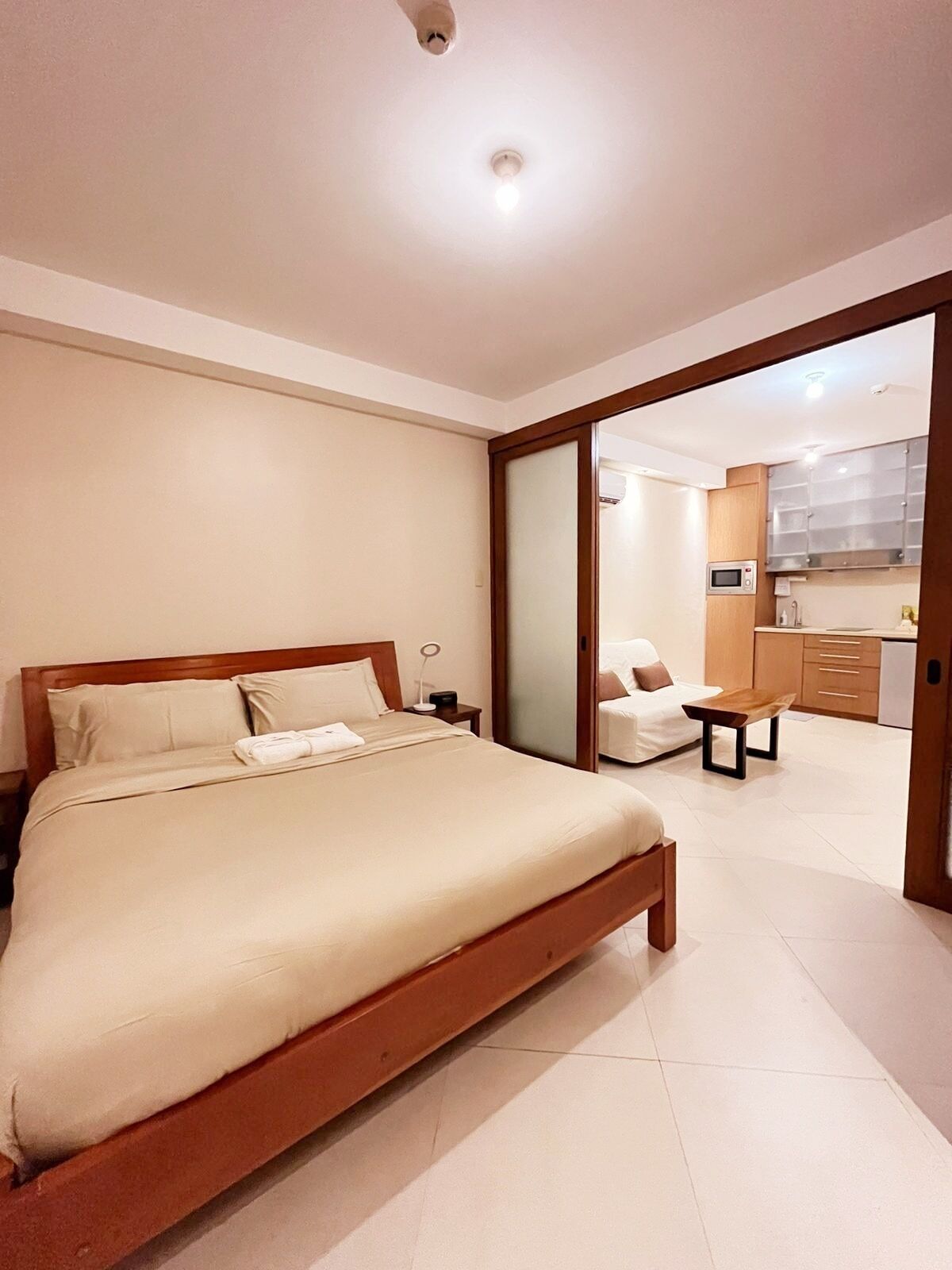 A bedroom with a bed, bedside table, and a view into a kitchen area with cabinets and a microwave.