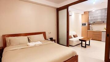A bedroom with a bed, bedside table, and a view into a kitchen area with cabinets and a microwave.