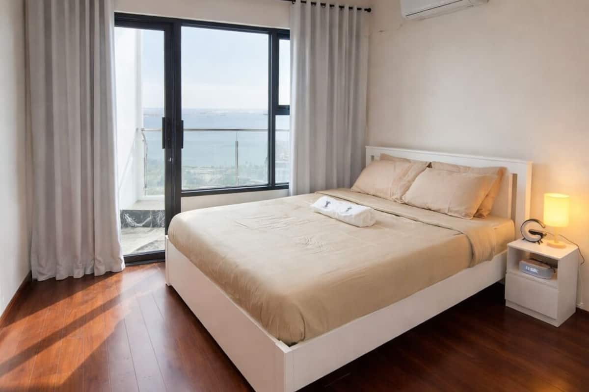 A bedroom with a bed, a nightstand, a lamp, a window with curtains, and a view of the outside.