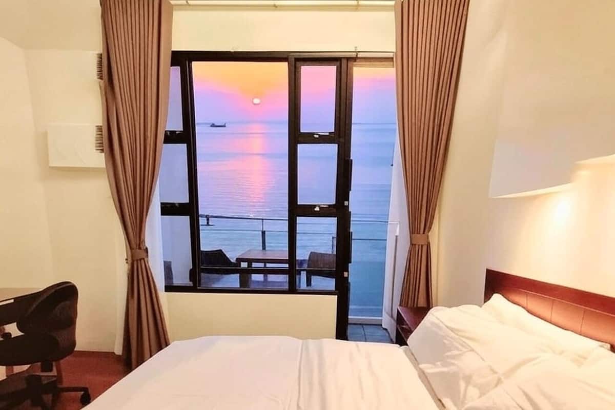 A hotel room with a bed, a desk, and a view of the sea at sunset.