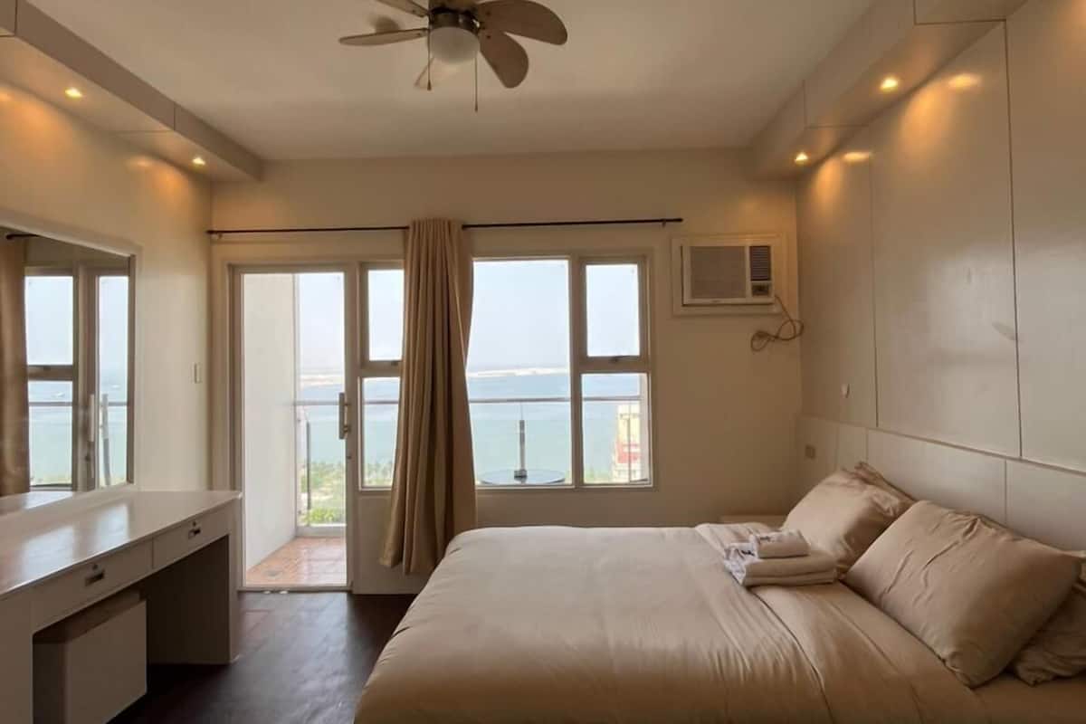 A bedroom with a bed, a desk, a ceiling fan, a window with a view, and an air conditioning unit.