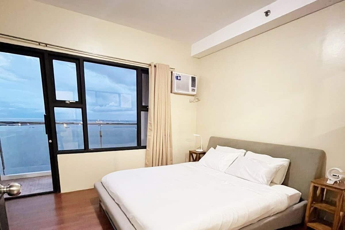 A bedroom with a bed, a nightstand, an air conditioning unit, and a view of the water through the window.