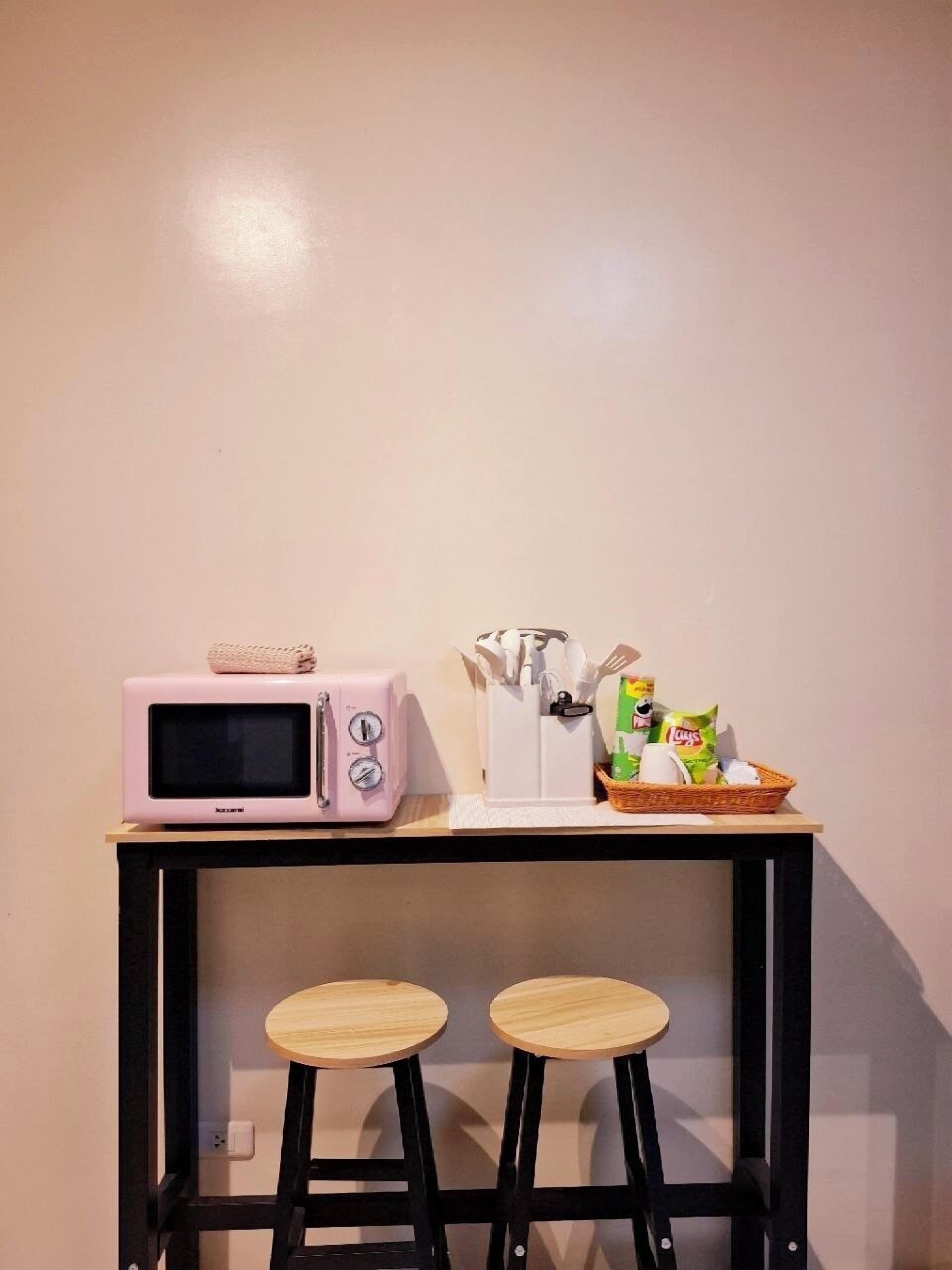 A kitchenette with a pink microwave, a white container with utensils, and a basket with snacks. There are two stools under a black table.
