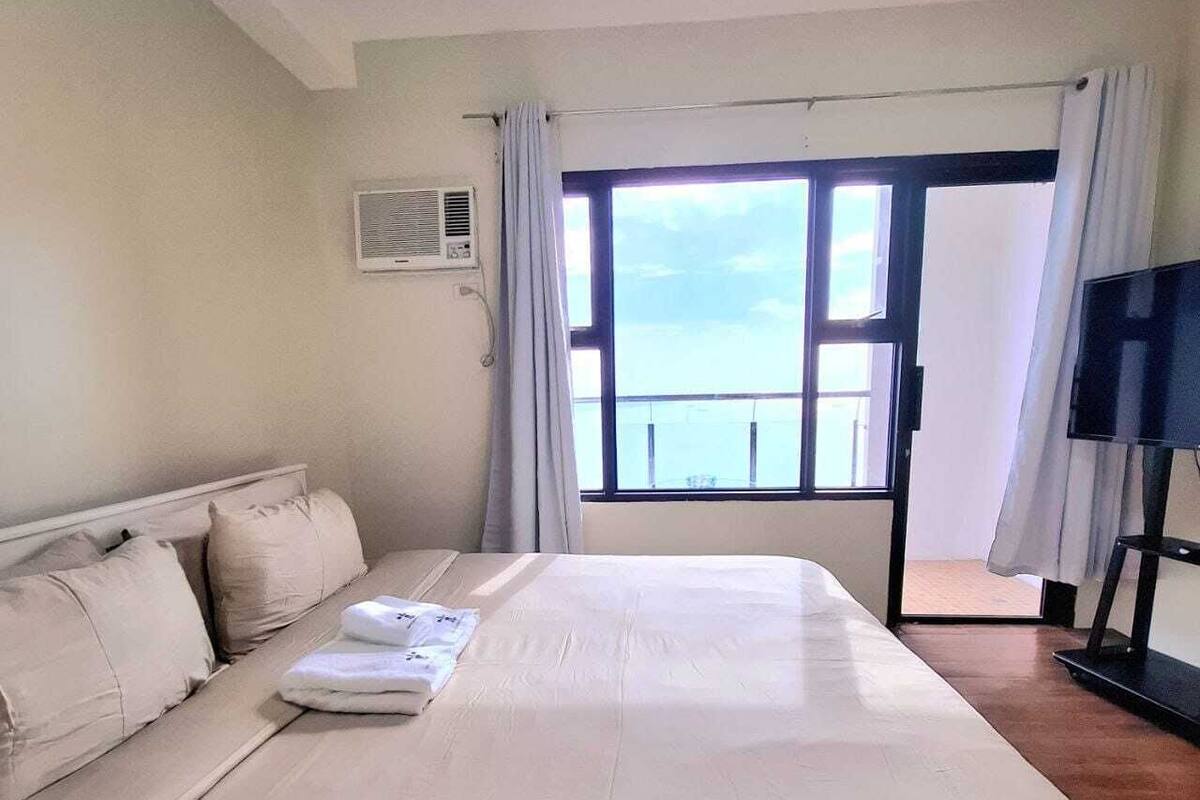 A hotel room with a large bed, a TV, a window with curtains, and a view of the sea.