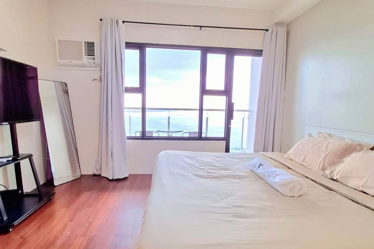 A hotel room with a large bed, a TV, and a view of the sea through a large window.