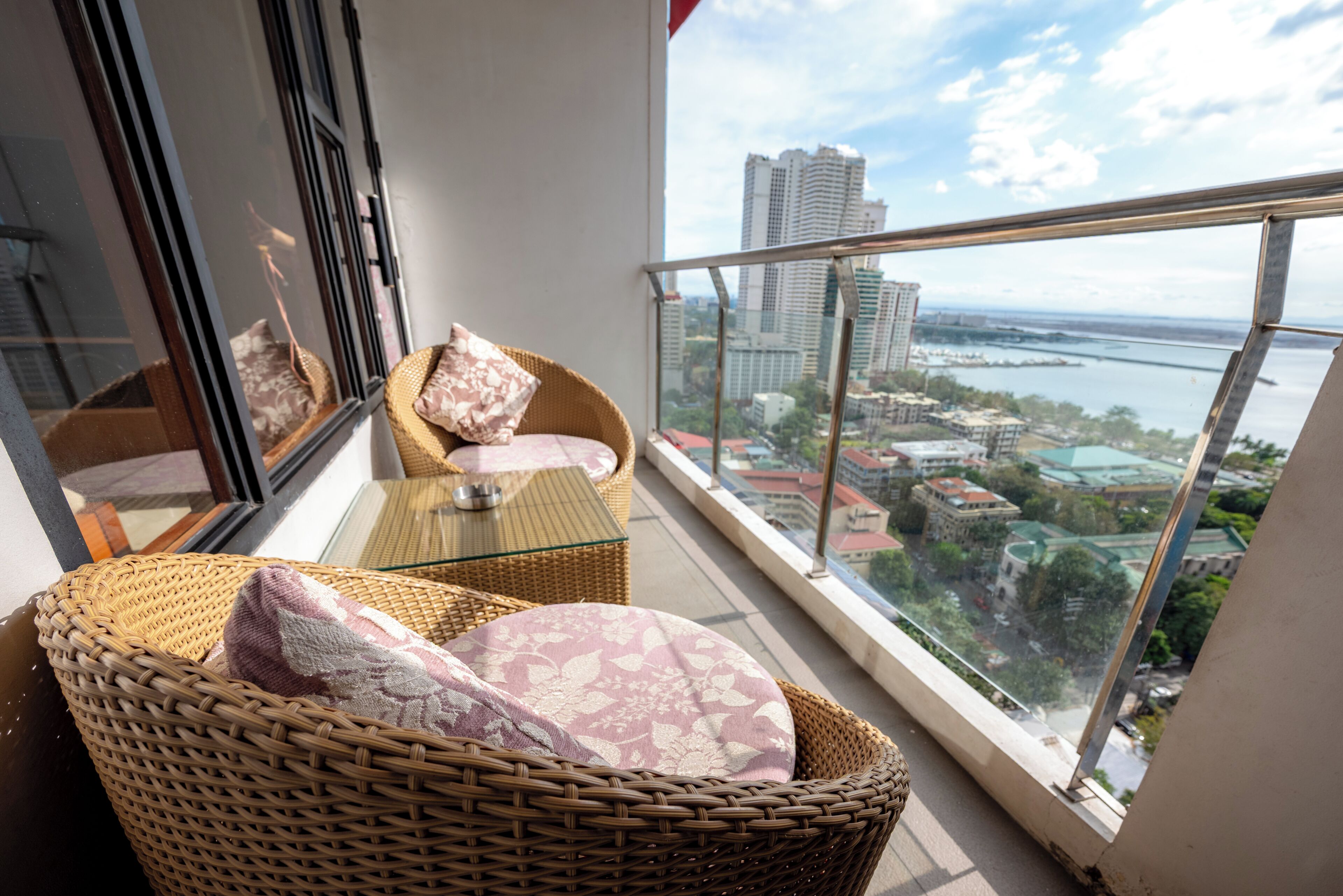 A balcony with wicker furniture, a glass table, and a view of a cityscape and water.