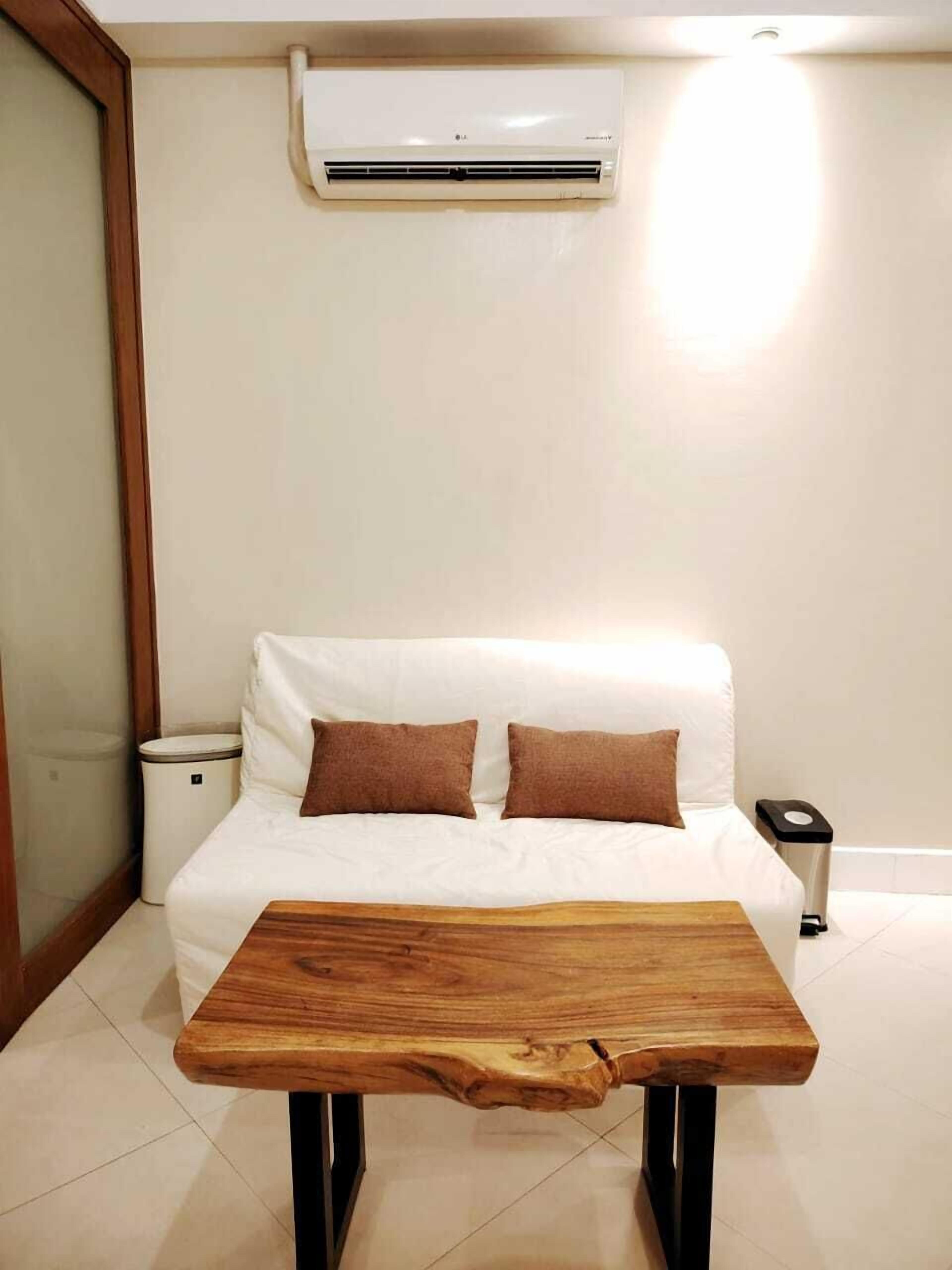 A bedroom with a white bed, two brown pillows, a wooden table, a white trash bin, and a white air conditioning unit on the wall.