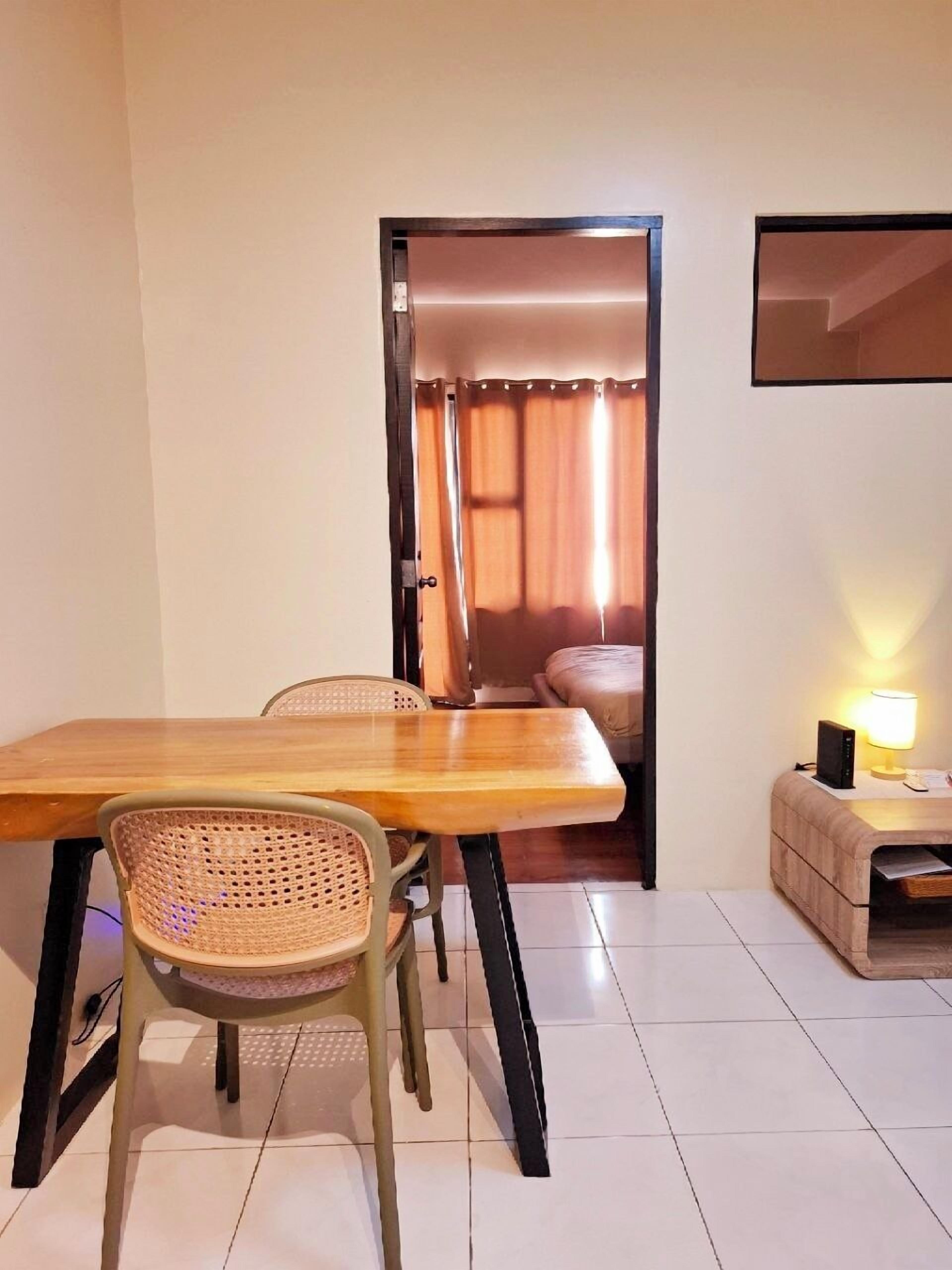 Una stanza con un tavolo da pranzo in legno, due sedie in vimini, un piccolo comodino con una lampada e una porta che dà su una camera da letto con un letto e tende.